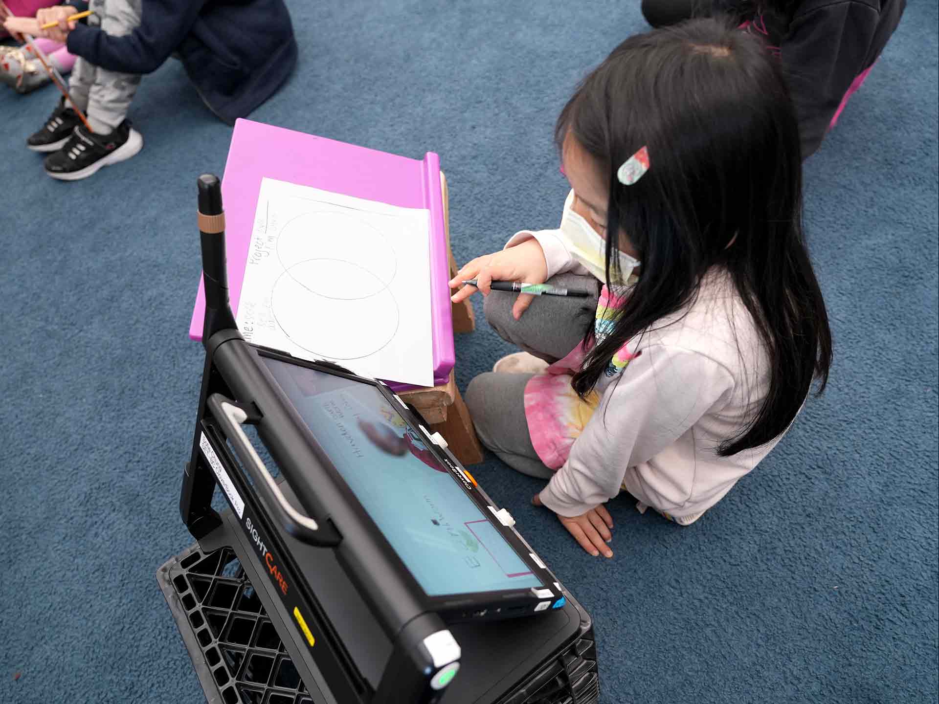 Sofie, a low vision student, working on her CloverBook and a writing board in the classroom at carpet time.