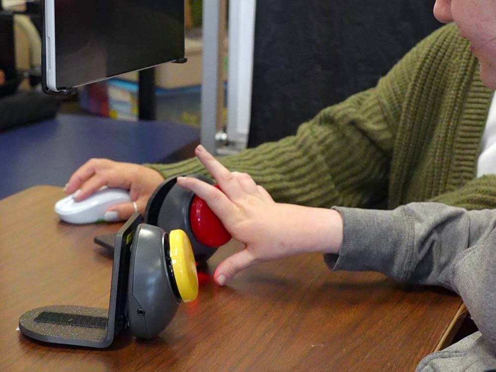 Student hand landing on a red assistive switch mounted on a desk in front of a him/her, along side with a yellow one.
